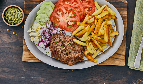 Fresh, tasty and delicious beef burger meat with French fries, tomato, garlic, onion, cucumber, pepper, Spanish onion,  pumpkin seeds and chia seeds, served  on a plate, on a dark wooden bar, restaurant or home kitchen table, close up view with copy space, representing fast food and city life, indulgence and joy through gourmet lifestyle