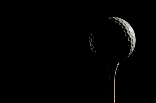 Golf ball and white tee with strong sidelighting against a black background and with room for copy left.