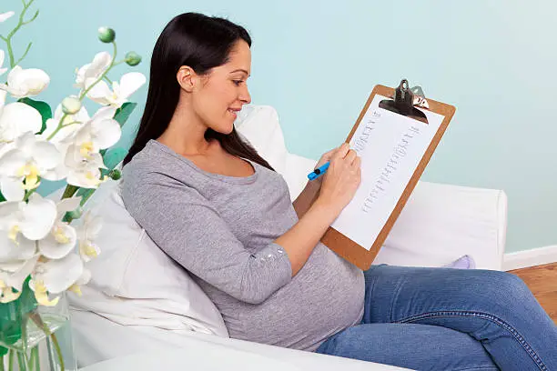 Photo of a pregnant woman at home sitting in an armchair writing a list of possible boy and girl names for her baby.