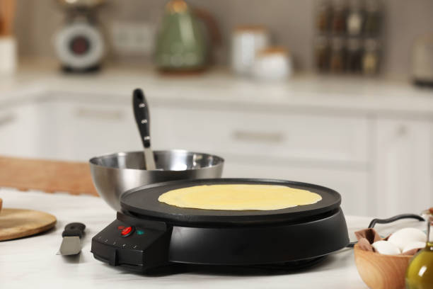 istock Cooking delicious crepe on electric pancake maker on white marble table in kitchen, space for text 1771266541