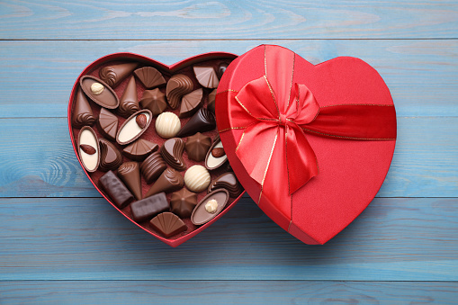 Heart shaped box with delicious chocolate candies on light blue wooden table, top view