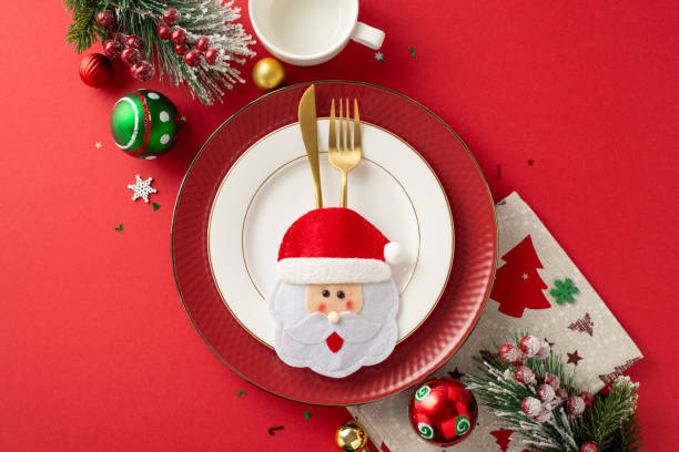 istock Vibrant New Year's party table setup for kids. Top-down view featuring plates, whimsical pocket utensil holder, mug, napkin, baubles, snowflake confetti, frosty fir twigs, and mistletoe berries on red 1771352052