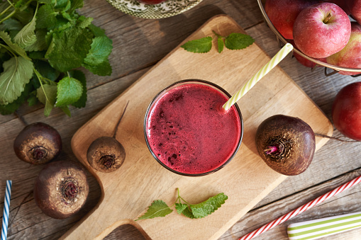 Cold drink made of beet juice in blue glass. Fresh beet, whole and sliced, on cutting board. Various green herbs on the background. Spotty blue background. Close-up