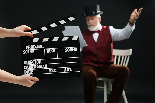 Senior actor performing role while second assistant camera holding clapperboard on black background, selective focus. Film industry