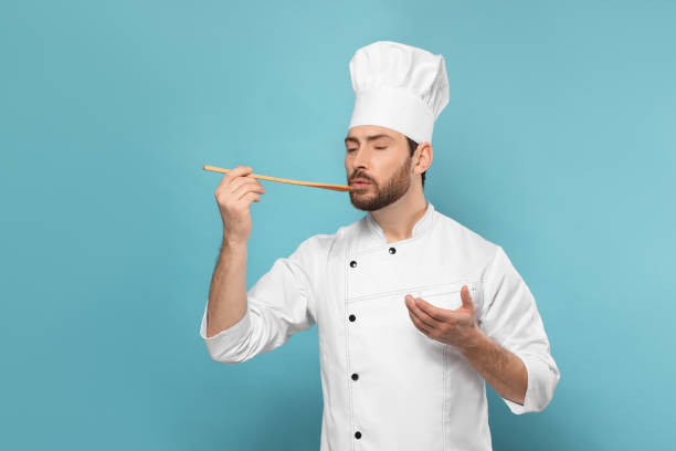 istock Mature male chef with spoon tasting dish on light blue background, space for text 1771390629