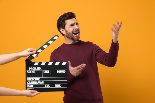 Emotional actor performing while second assistant camera holding clapperboard on orange background