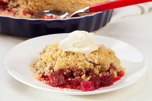 "A plate of rhubarb crumble with a dollop of cream, serving dish and spoon in the background."