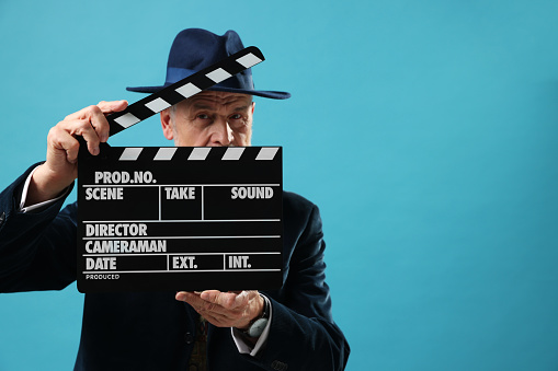 Senior actor holding clapperboard on light blue background, space for text. Film industry