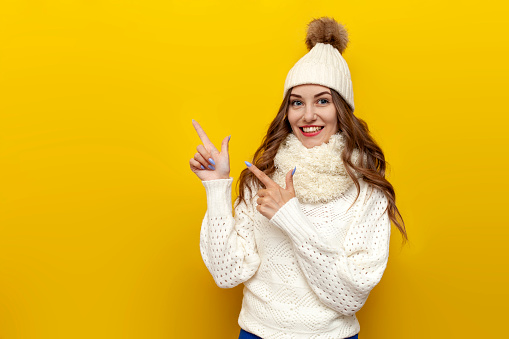 young cute girl in warm soft winter clothes points with her hands to the side on yellow isolated background, woman in white hat and scarf in comfortable and cozy knitted sweater advertises copy space