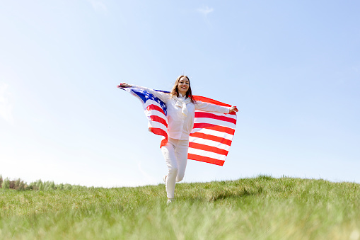 young girl runs through the field with the flag of america and smiles, the concept of freedom and the fourth of july