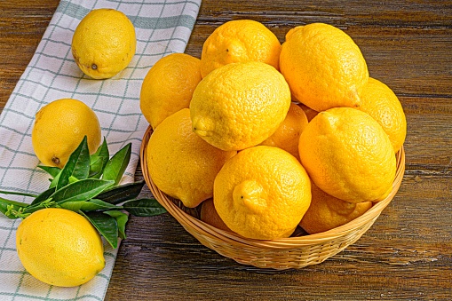 A white ceramic basket filled with freshly-harvested lemons is placed on top of a rustic wooden table