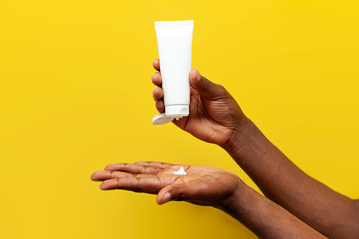 hands of african american man hold white empty tube of cream on yellow isolated background, hands squeeze out skin care cream, close-up