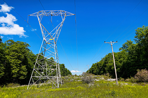Two types of electric towers are fading off into the distance  on a green easement right of way through the forest of Garret Maryland. The sky is blue with a few clouds. The electric cables run from the top of the image through the middle of the image.