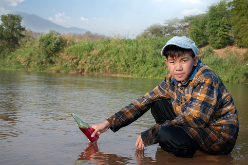 Asian boy in plaid shirt and cap holds bottle which has EM or Effective Microorganisms to study water, river,dirt, chemical, tiny underwater insects and underwater plant in the river nearby his village.