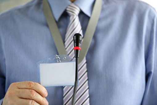 Guy in business clothes in front of a microphone shows a badge. Employee shows a document in the audience