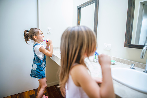 Side view close-up of 6 and 8 year old Hispanic sisters standing at bathroom sinks and brushing their teeth.
