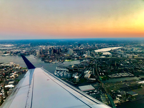 Embraer airplane flying over Boston city in Massachusetts. The river is visible and skyscrapers on the ground. The sky is clear and the sunset is a mix of blue, orange, yellow, and pink.