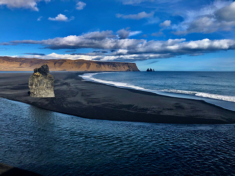 A tall cliff behind a black sand beach next to the Atlantic Ocean in Iceland. There are small islands in the background. The waves are crashing onto the shore. The sky is blue with clouds.
