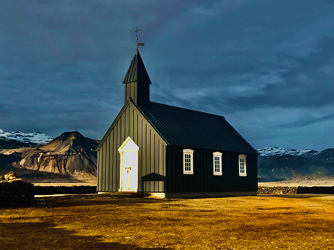 Famous black church in front of mountains with snow-capped peaks in a clearing with yellow grass in Iceland. The church has white doors and windows. The sky is dark and cloudy.