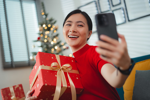 Asian woman enjoying using smartphone for video calling or live streaming vlog showing gifts on Christmas Day, sitting in living room at home.