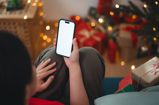 Over the shoulder shot of woman holding smartphone with white screen sitting on sofa in living room decorated with a Christmas tree.