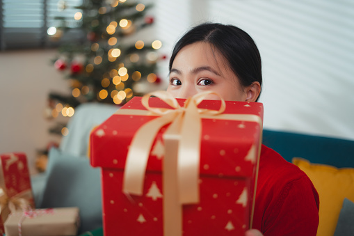 Asian woman happy excited holding gift box for Christmas in sitting in living room decorated with a Christmas tree and looking at camera.
