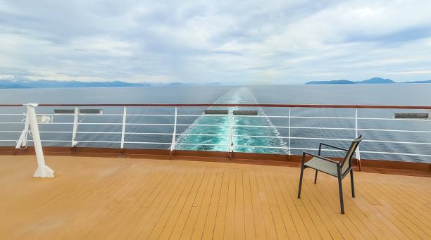 An empty deck chair on the fantail, or stern, of a cruise ship.   The wake of the large ship is visible and stretches to the horizon.   The water is green with minerals from the surrounding glacier runoff.  The seat beckons the viewer to be in the seat; to feel the cool breeze and see the beautiful scenery around them.  The image has a quiet, tranquil feel to it.  The photo was taken on an Alaskan Inside Passage cruise.