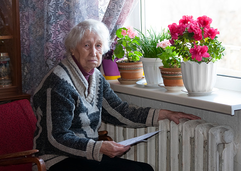 The senior woman holding gas bill in front of heating radiator. Payment for heating in winter.