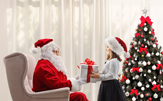 Santa claus sitting in an armchair and giving a present to a girl next to a christmas tree