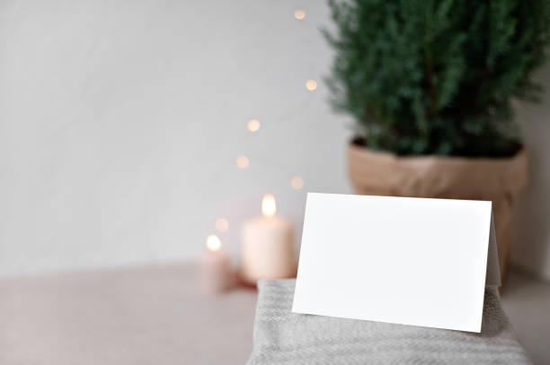 istock Blank paper card mockup, sweaters on table, blurred defocused candle and garland lights and juniper in pot on background. Empty neutral beige wall copy space. Christmas postcard or invitation template 1772264920