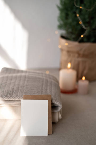 istock Blank paper card mockup, envelope, knitted sweaters on table, blurred candle and garland lights on background. Natural sunlight shadow. Christmas, New Year winter home decor, postcard template 1772264974