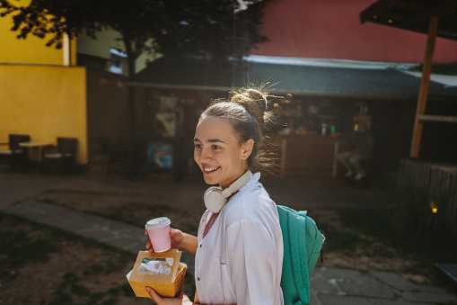 Photo of a female healthcare professional going to work for a shift in a hospital. She is in her medical scrubs, equipped with a food and a coffee to go.