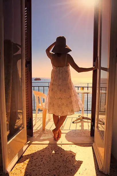 "A young woman on the balcony of a hotel room looking in the distance at Meditarrenean Sea with a ferry boat on it. This image was taken in Agia Marina (Aegina Island, Greece)"