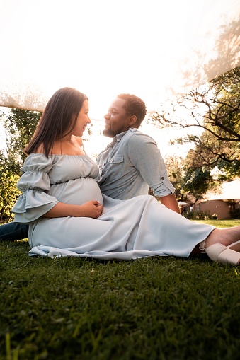A pregnant Latina woman and African man couple sitting in a lush green bright sunlit park