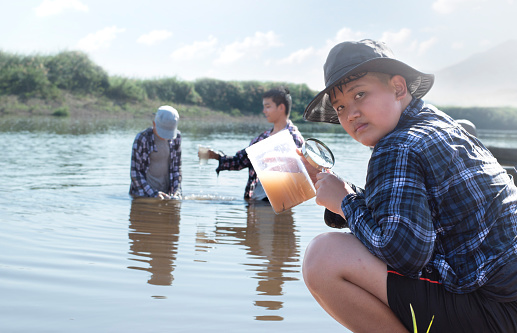 Asian schoolboys in plaid shirt and cap holding transparent tube which contained cloudwater and clearwater from local river to do outdoor science subject homework in topic 'climate change' concept.