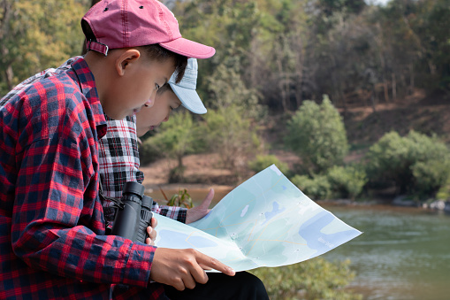 Asian boy in plaid shirt is taking his summer holiday at local national park with his friend by watching birds, insects, animals, trees, rivers, mountains and weather by using map, binoculars, laptop.