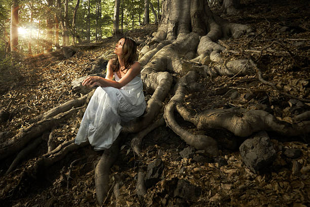 Lovely young lady wearing elegant white dress enjoying the beams of celestial light on her face sitting on the mighty roots of an ancient tree in enchanted woods