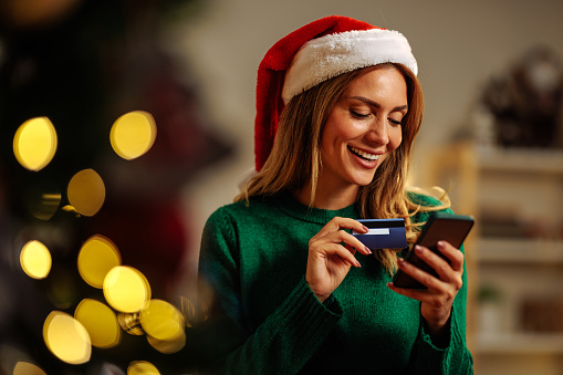 Woman wearing a Santa hat and holding a phone and a credit card while attempting to online buy a christmas present