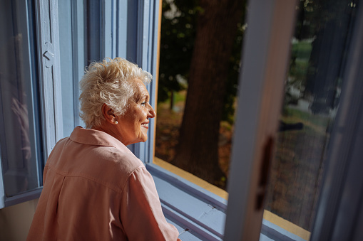 A view of a happy elderly woman standing in front of her blue window, enjoying her view and a breeze