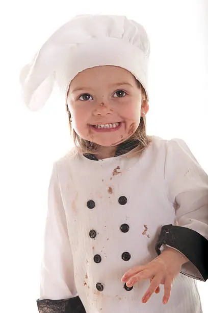 "An adorable preschooler in a chef's outfit, happily messy from her chocolate cake batter.  On a white background."