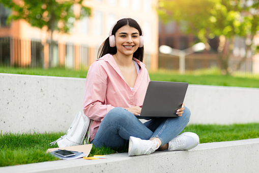 Happy hispanic student lady in wireless headphones studying with laptop outdoors, sitting with computer at campus, looking and smiling at camera, enjoying distance learning, copy space