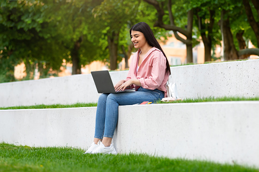 Happy teen hispanic lady with laptop sitting in park, student girl studying remotely with computer outdoors, typing on keyboard and smiling, copy space. Online learning concept