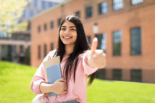Smiling hispanic student girl showing thumb up gesture and smiling at camera, standing in college campus, positive lady holding workbooks, recommending educational programs