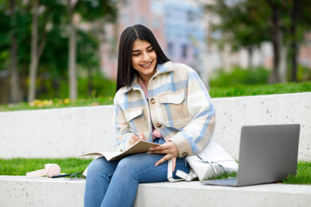 istock E-learning concept. Cheerful teen student girl learning outdoors, taking notes while sitting with laptop in park 1772367033