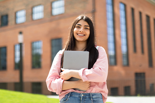 Portrait of happy hispanic lady student posing with laptop in hands outdoors, looking and smiling at camera, having break after classes in university campus