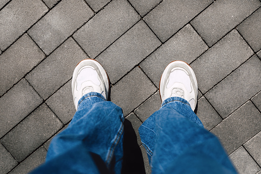 Women's legs in jeans and white modern sneakers on gray street tiles, copy space, top view and personal point of view