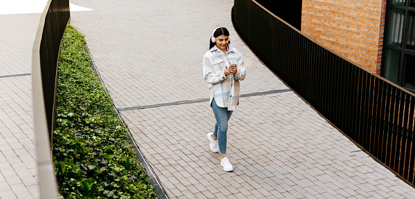 Happy teenage lady chatting in social networks on cellphone and wearing headphones while walking in urban area in the city, panorama, copy space