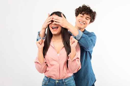 Cheerful young boyfriend surprising girlfriend covering eyes standing behind her back over white studio background. Unexpected romantic surprise, Valentine's day celebration