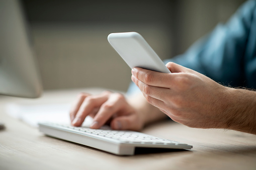 Unrecognizable businessman wprking with smartphone and computer in office, male hands holding mobile phone and typing on pc keyboard, using modern gadgets for business, cropped shot, closeup