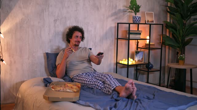 Young Man Eating Pizza And Watching TV In The Bed At Night